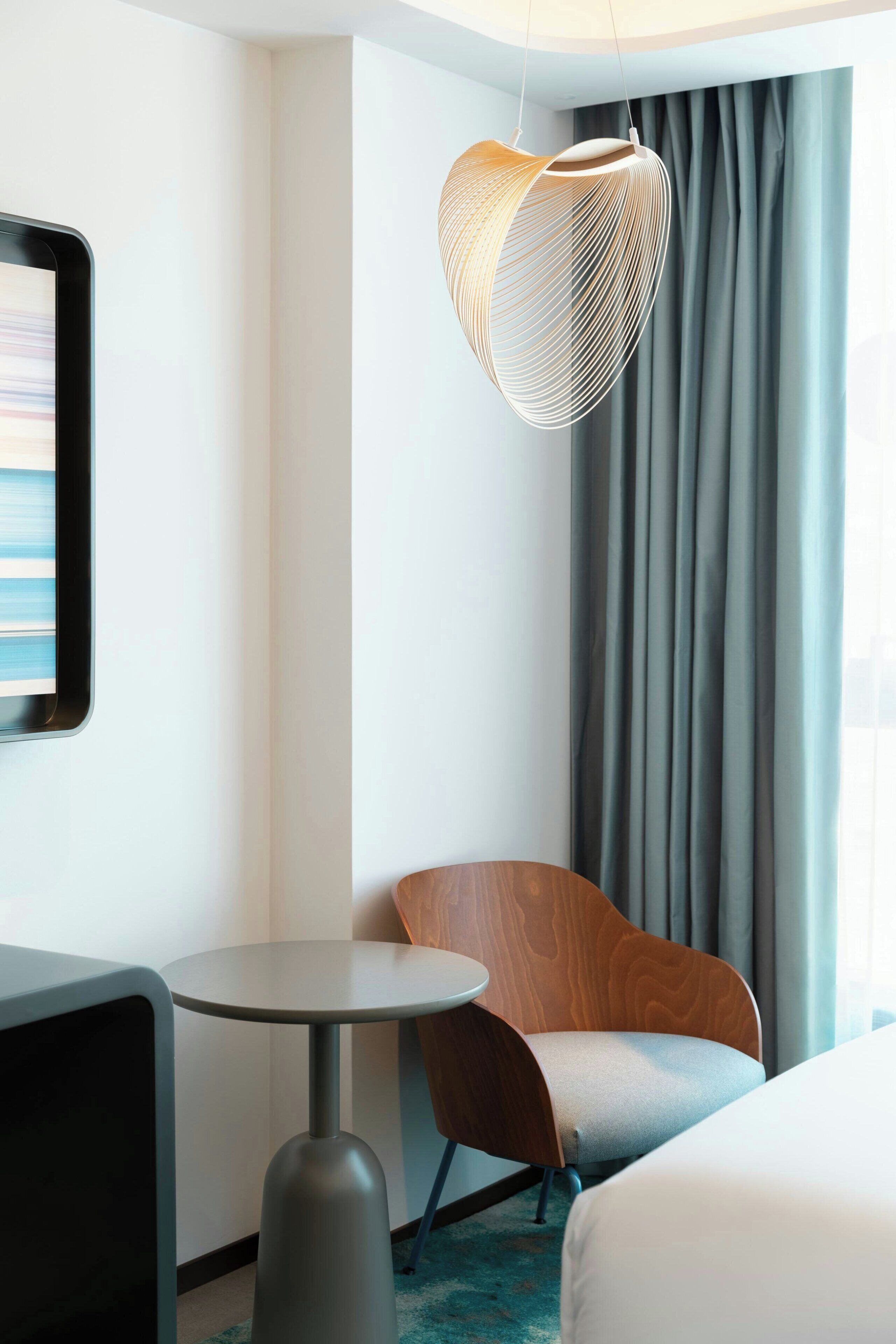 A modern hotel room with a bed, a round table, a wooden chair, and a unique hanging light fixture.