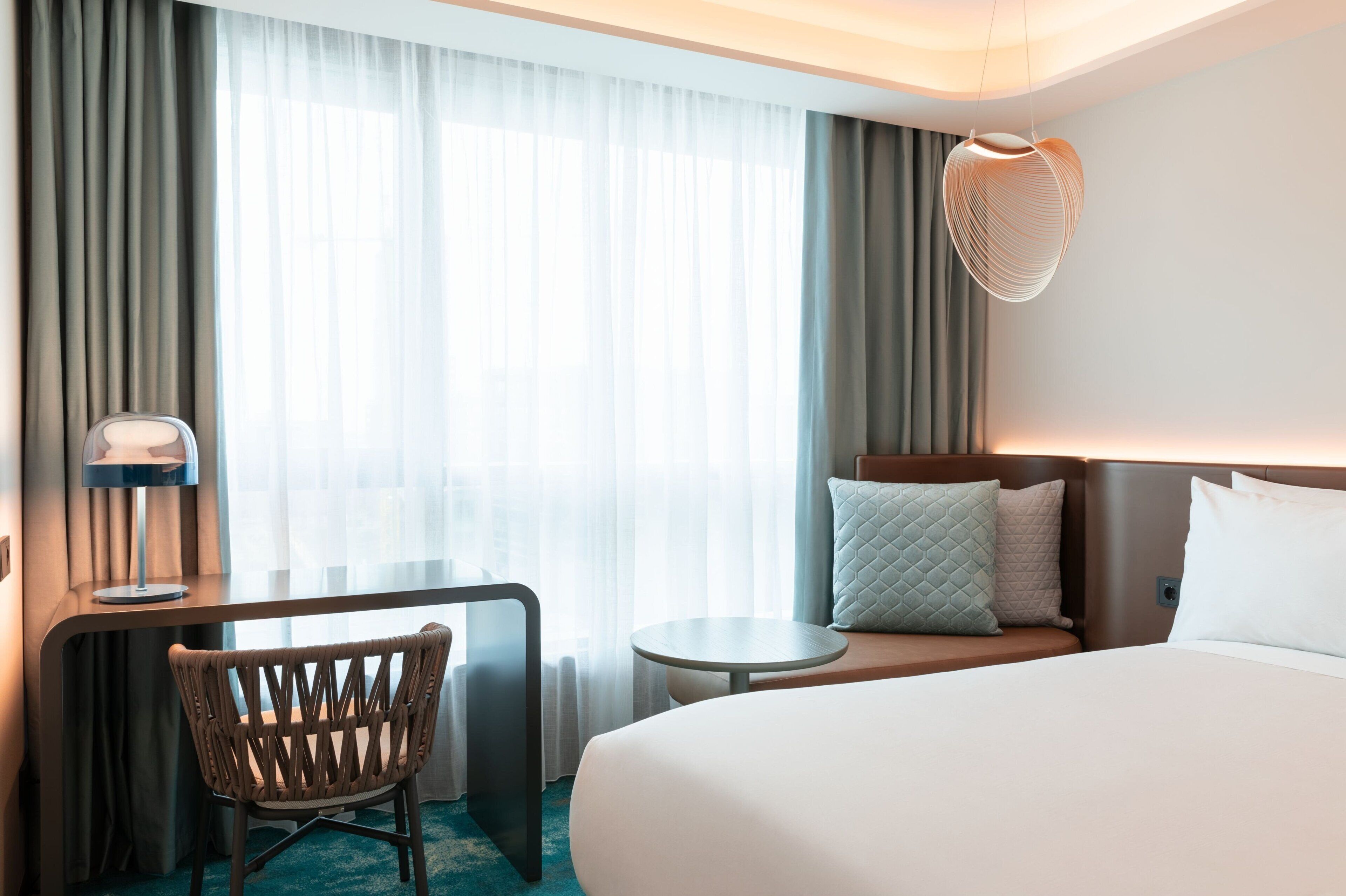 Une chambre d’hôtel comprenant un lit, un bureau avec une chaise, une petite table et un luminaire suspendu.