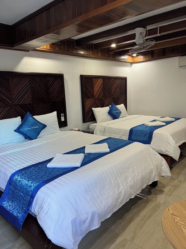 A hotel room with three beds, each with white bedding and blue accents, wooden headboards, and a ceiling fan.