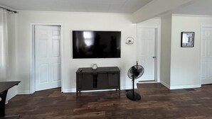 Een woonkamer met een flatscreen televisie aan de wand, een donker houten kast, een vloerventilator en een deur naar een andere kamer.