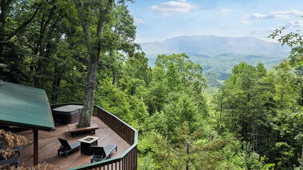 A wooden deck with outdoor furniture, surrounded by dense forest and a mountain view. - 4BD Rustic Log Cabin With Incredible Deck and View! (Gatlinburg)
