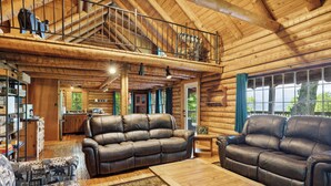 A spacious log cabin interior with a high ceiling, leather sofas, a wooden coffee table, and a balcony overlooking a scenic view. - 4BD Rustic Log Cabin With Incredible Deck and View! (Gatlinburg)