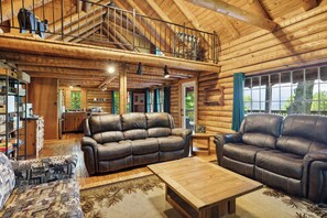 A spacious log cabin interior with a high ceiling, leather sofas, a wooden coffee table, and a balcony overlooking a scenic view. - 4BD Rustic Log Cabin With Incredible Deck and View! (Gatlinburg)