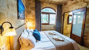 A bedroom with stone walls, a bed with white linens, a night light, and a window with curtains. - Villa Soldanis Sleeps 12 with Pool (Xaghra)