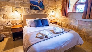 A cozy room with a stone wall, a bed with white linens, two pillows, a nightstand, and a painting of a coastal scene. - Villa Soldanis Sleeps 12 with Pool (Xaghra)