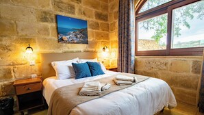 A neatly made bed with white and blue linens, a stone wall, and a painting of a coastal landscape. - Villa Soldanis Sleeps 12 with Pool (Xaghra)
