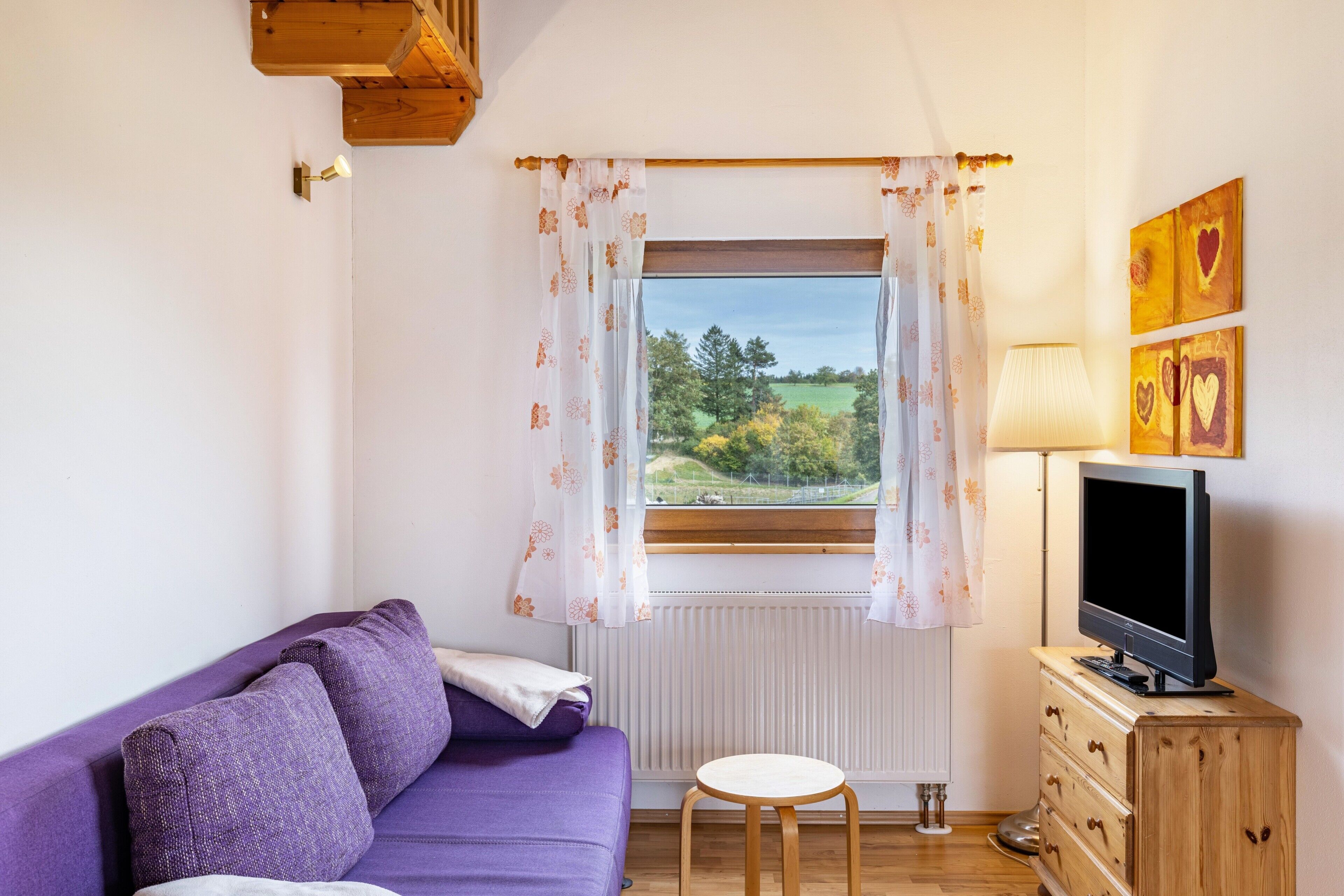 Ein Zimmer mit einem violetten Sofa, einem Fernseher auf einer Holzkommode, einem Fenster mit Vorhängen und einer Heizung.