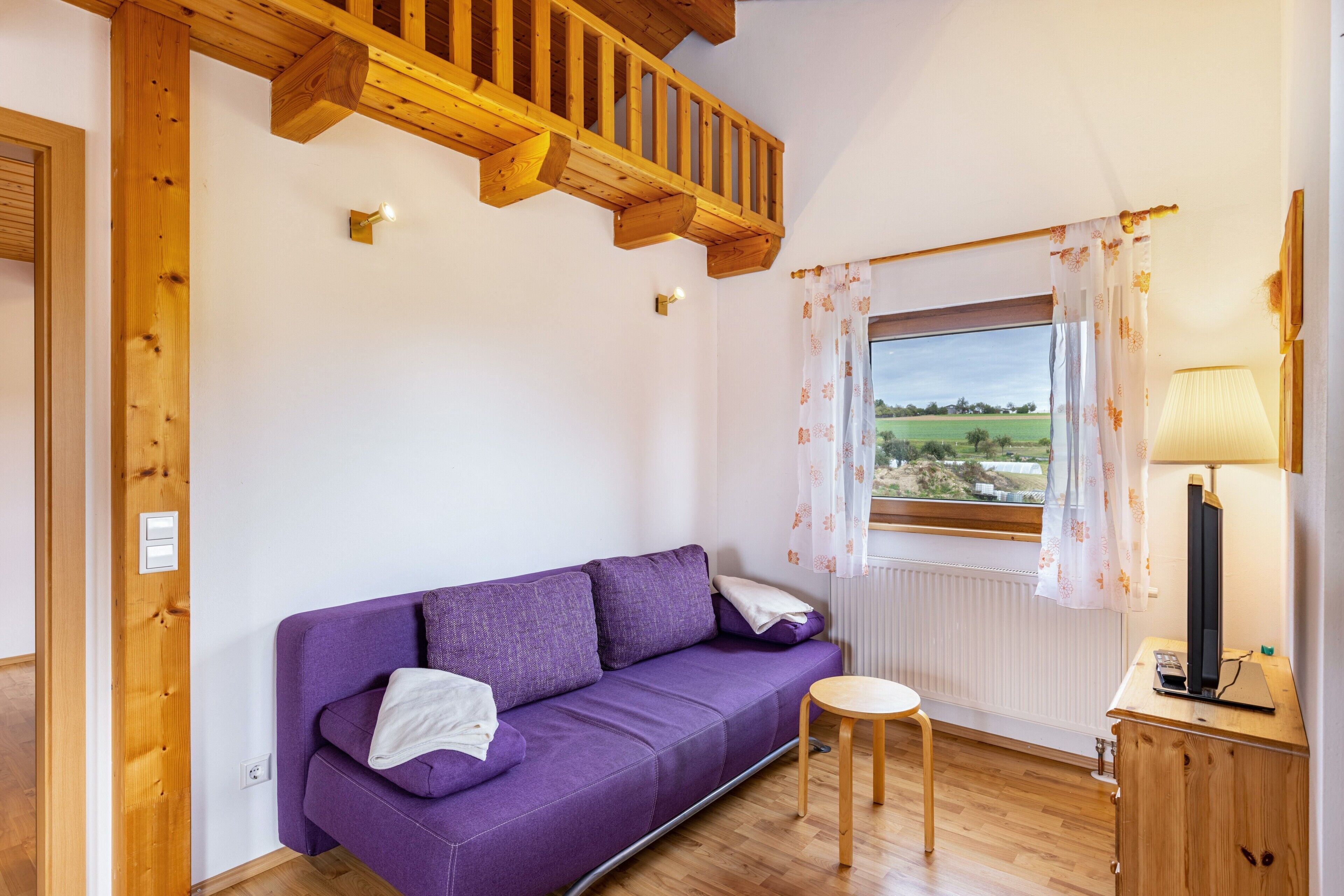 Una habitación con un sofá púrpura, una pequeña mesa de madera, una lámpara y una ventana con vistas al campo.