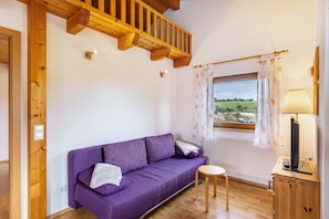 Une pièce avec un canapé violet, une petite table en bois, une lampe et une fenêtre donnant sur la campagne.