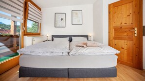Une chambre à coucher avec un lit, deux tableaux encadrés au mur, une porte en bois et une fenêtre avec des stores.