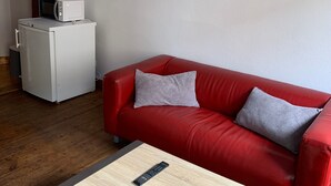 En röd soffa med två kuddar, ett litet bord med en fjärrkontroll och en växt, ett vitt kylskåp och en tavla på väggen.