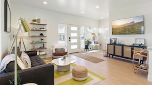 A spacious living room with a sofa, coffee table, and ottoman, a bookshelf, a desk with a computer, and a large window with a view of a field. - Modern Downtown Talent Dream (Talent)