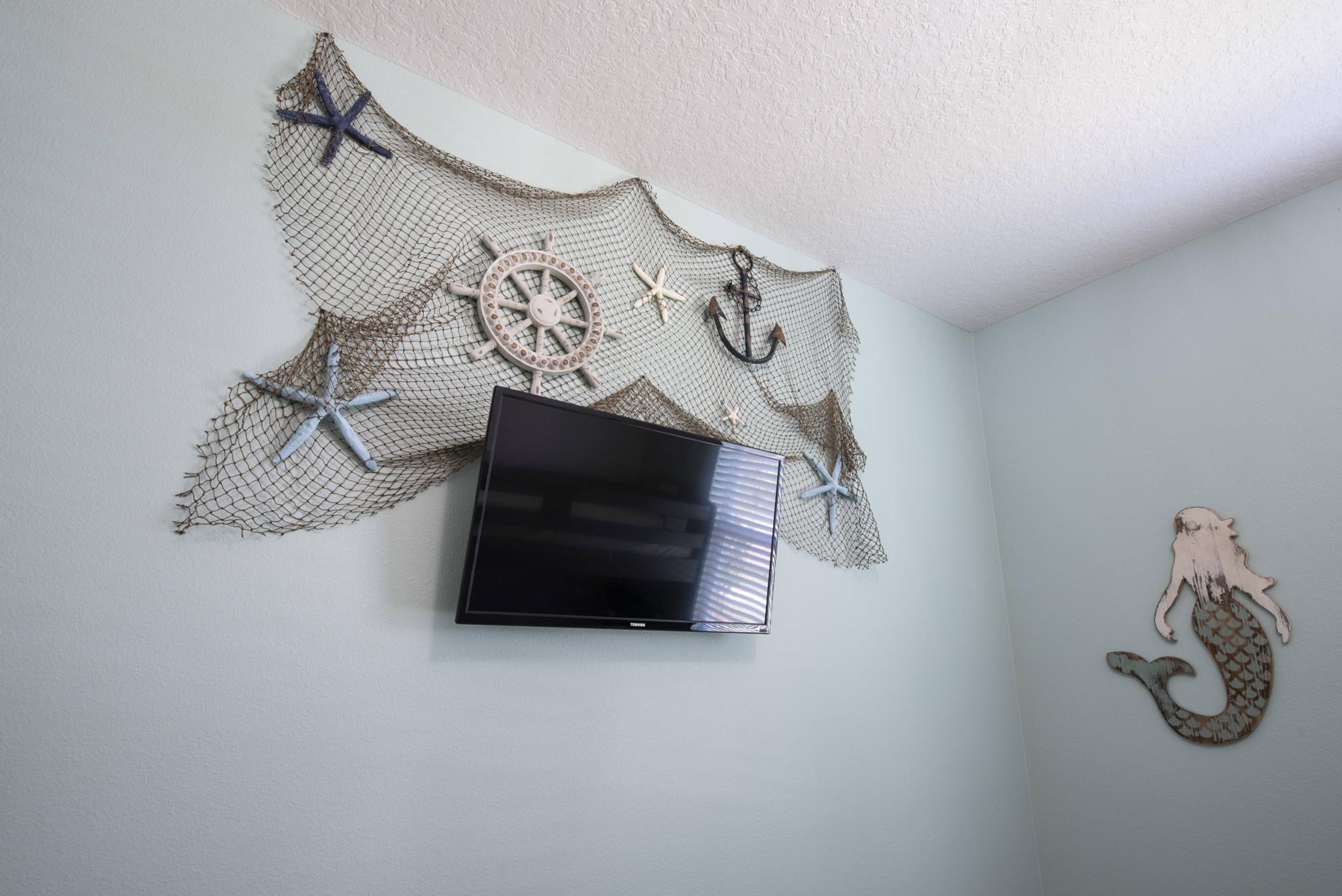 A flat-screen TV mounted on a wall with nautical-themed decorations including a ship's wheel, anchors, and a mermaid figure.