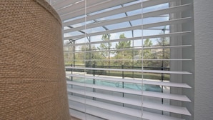 Ein Zimmer mit weißen Jalousien, Blick auf einen Pool und ein Korbstuhl.