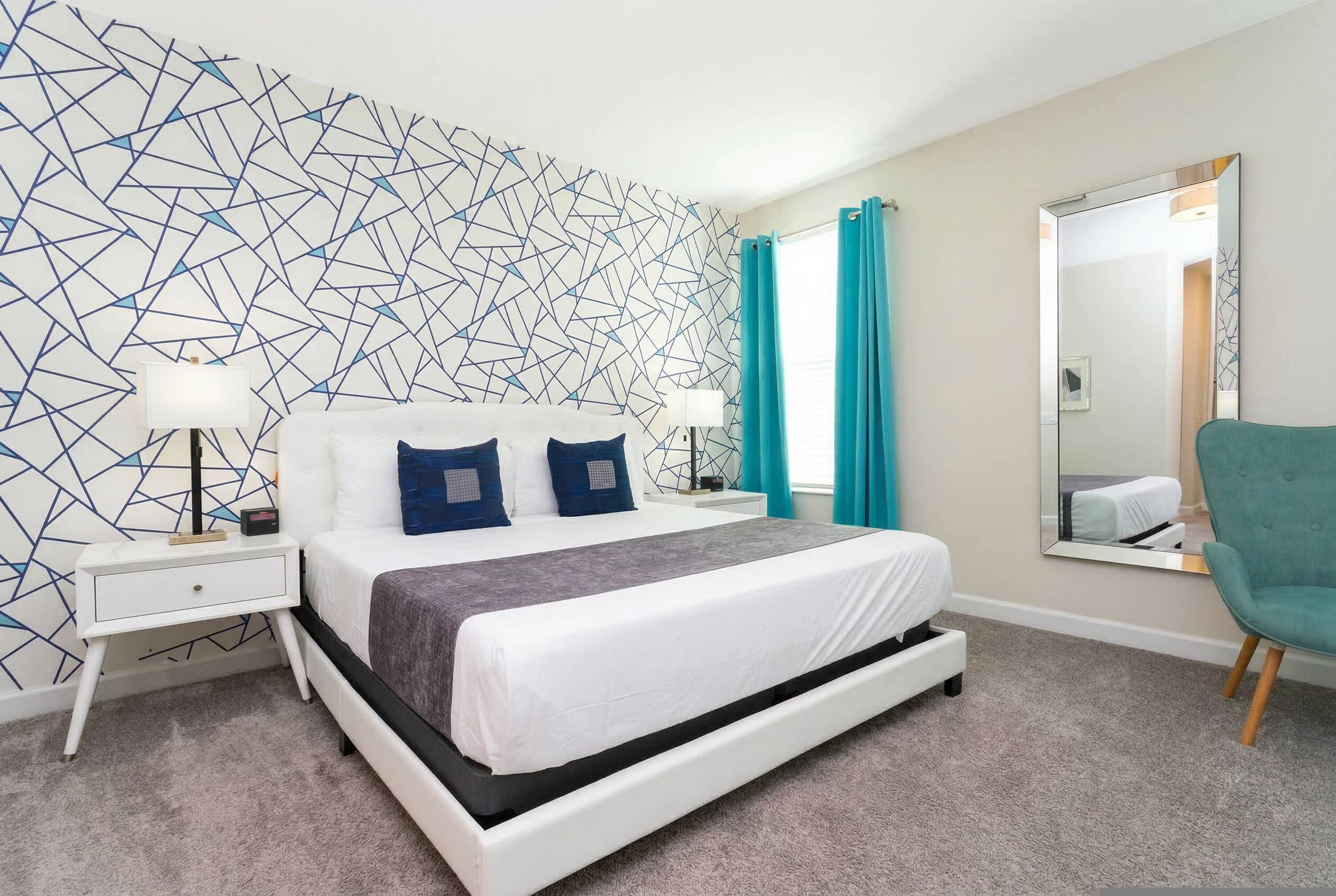 A modern hotel room with a geometric-patterned wall, a white bed with gray and blue bedding, a teal armchair, and a large mirror.