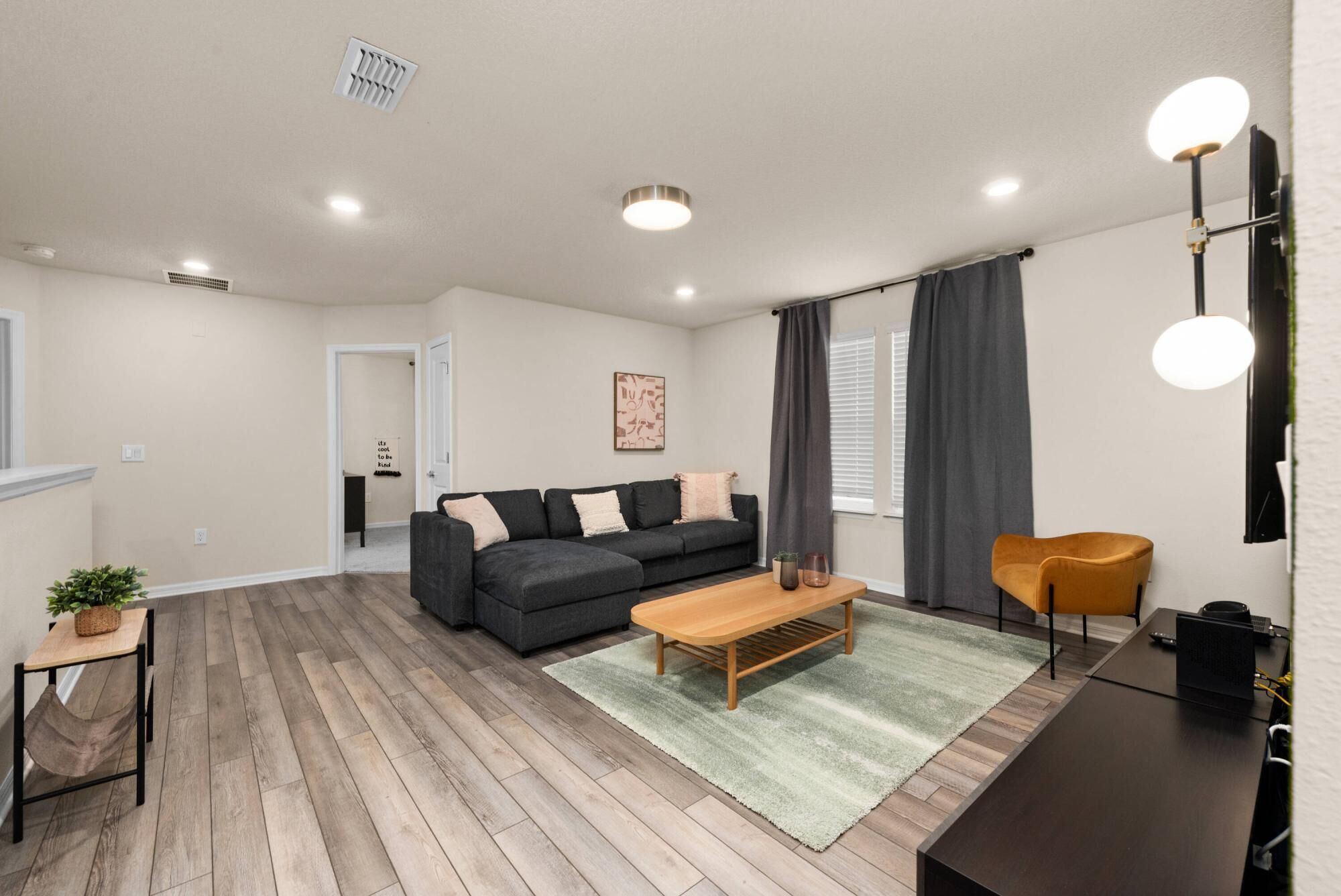 Ein modernes Wohnzimmer mit einem dunkelgrauen Sofa, einem Holztisch und einem gelben Sessel. Ein kleiner Beistelltisch mit Pflanze und Lampe steht daneben. Der Raum hat Holzböden und ein großes Fenster mit grauen Vorhängen.