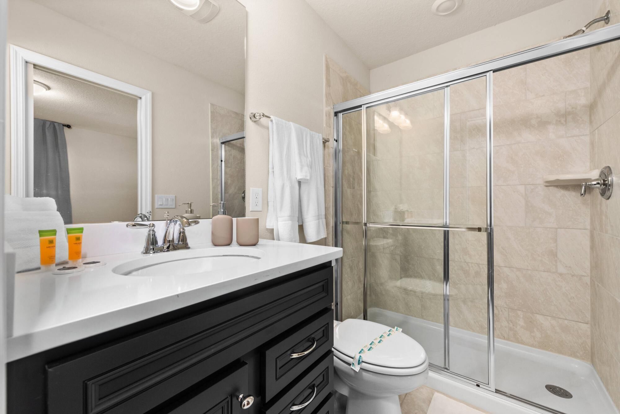 A modern bathroom with a glass-enclosed shower, a white sink with two bottles of shampoo, and a black vanity with drawers.
