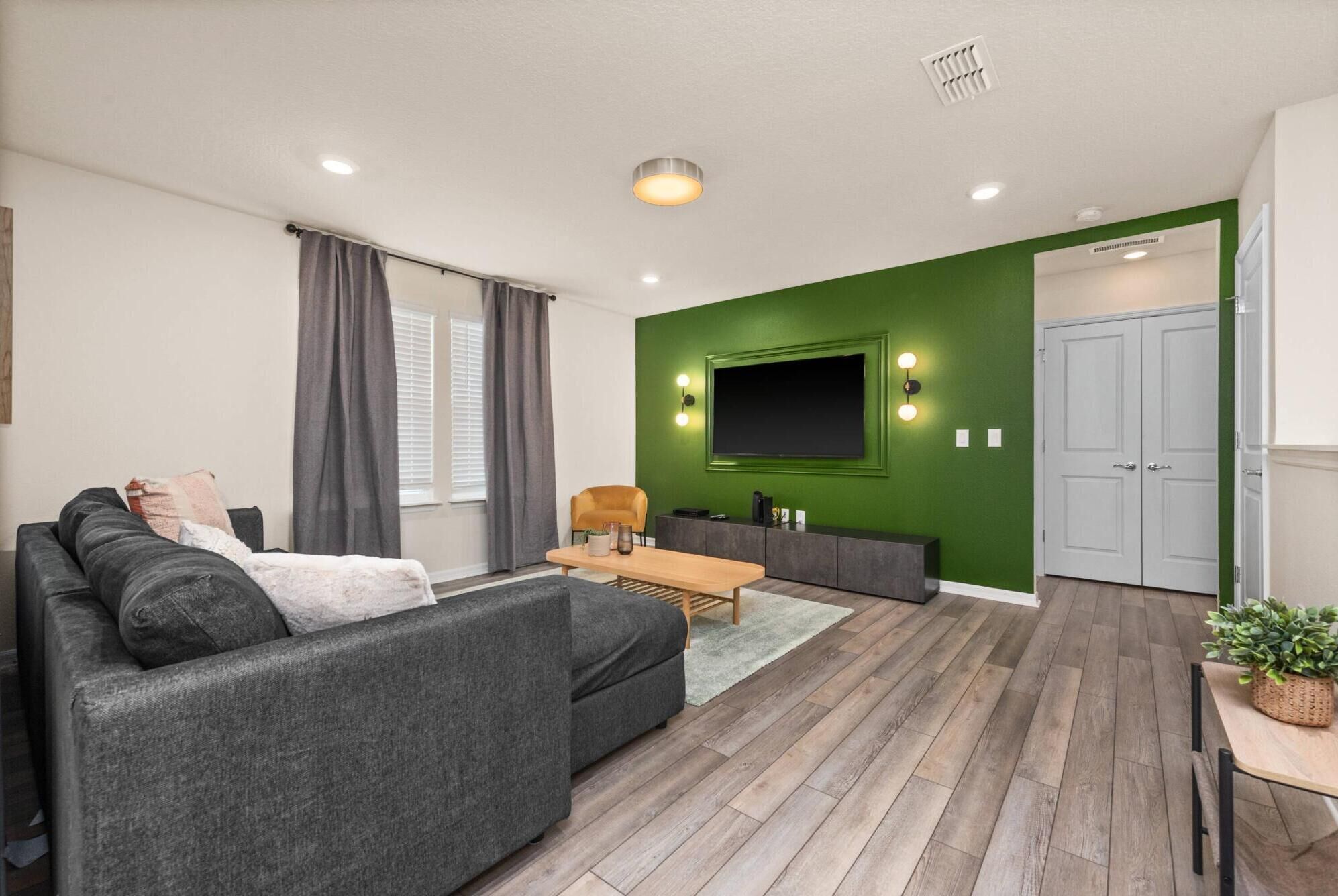 A modern living room with a grey sofa, a coffee table, and a TV mounted on a green wall. There is a door and a window with curtains.