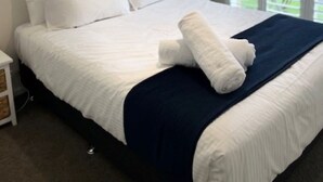 Un lit bien fait, avec du linge de lit blanc et une couverture bleu marine, un tableau encadré au mur et une fenêtre avec des stores.