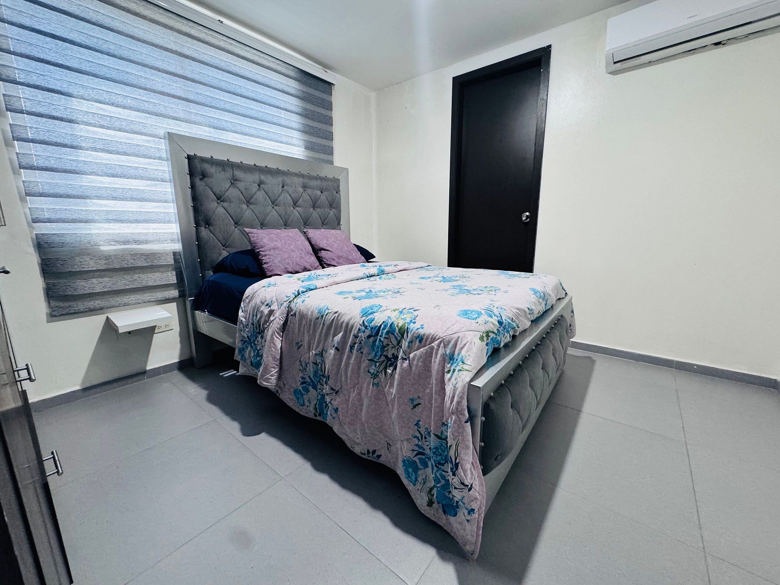 Un dormitorio con cama, ventana con persianas, una puerta y un equipo de aire acondicionado.
