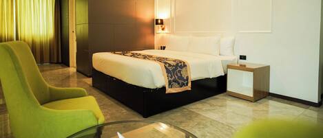 Una habitación de hotel moderna con una cama grande, una mesita de vidrio, un sillón verde y una mesita de noche con lámpara.