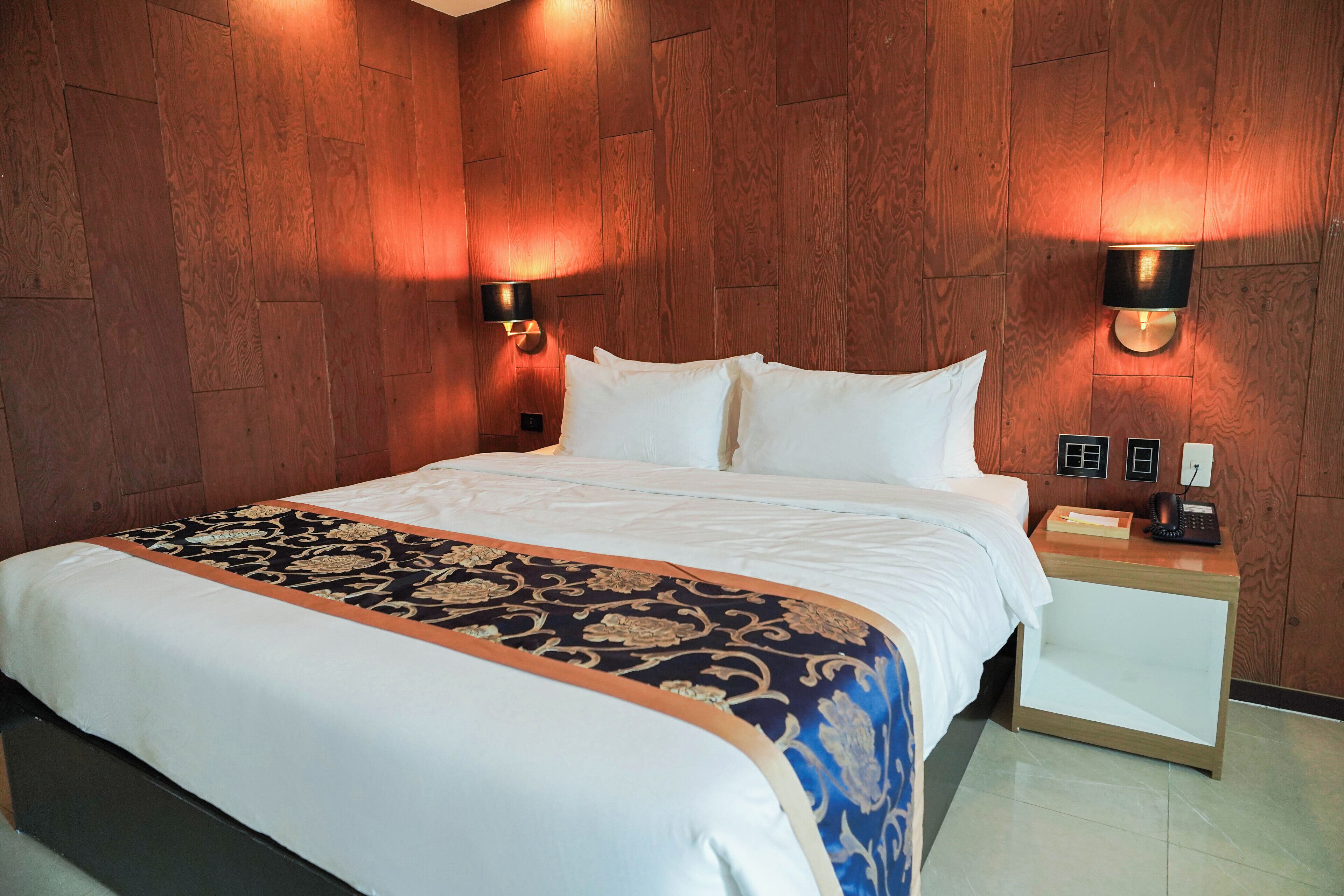 Habitación de hotel con una cama grande, paneles de madera en las paredes, dos lámparas fijadas a la pared, una mesita de noche con teléfono y una mesita auxiliar.