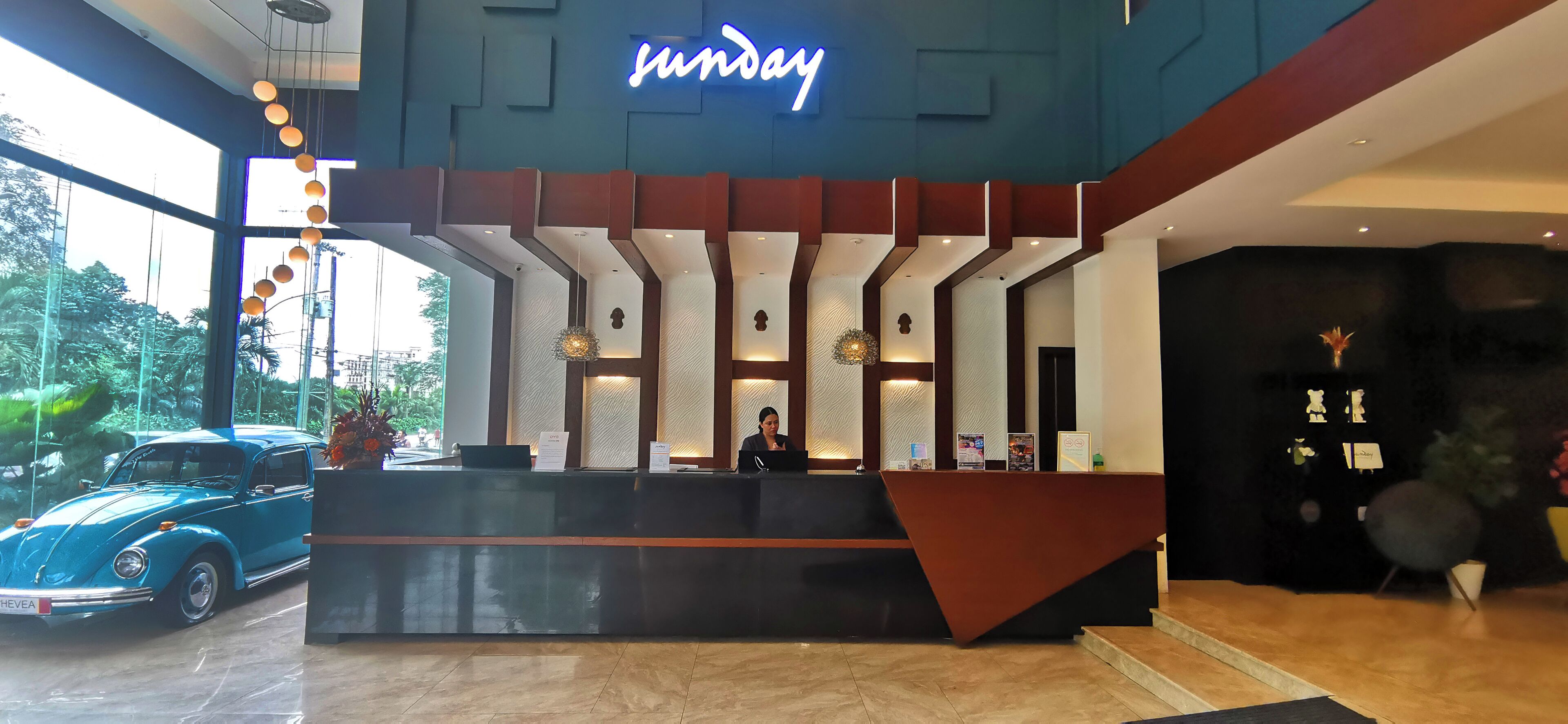 A hotel reception desk with a neon sign, a receptionist, and a car parked outside.