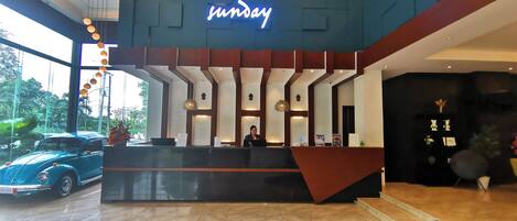 A hotel reception desk with a neon sign, a receptionist, and a car parked outside.