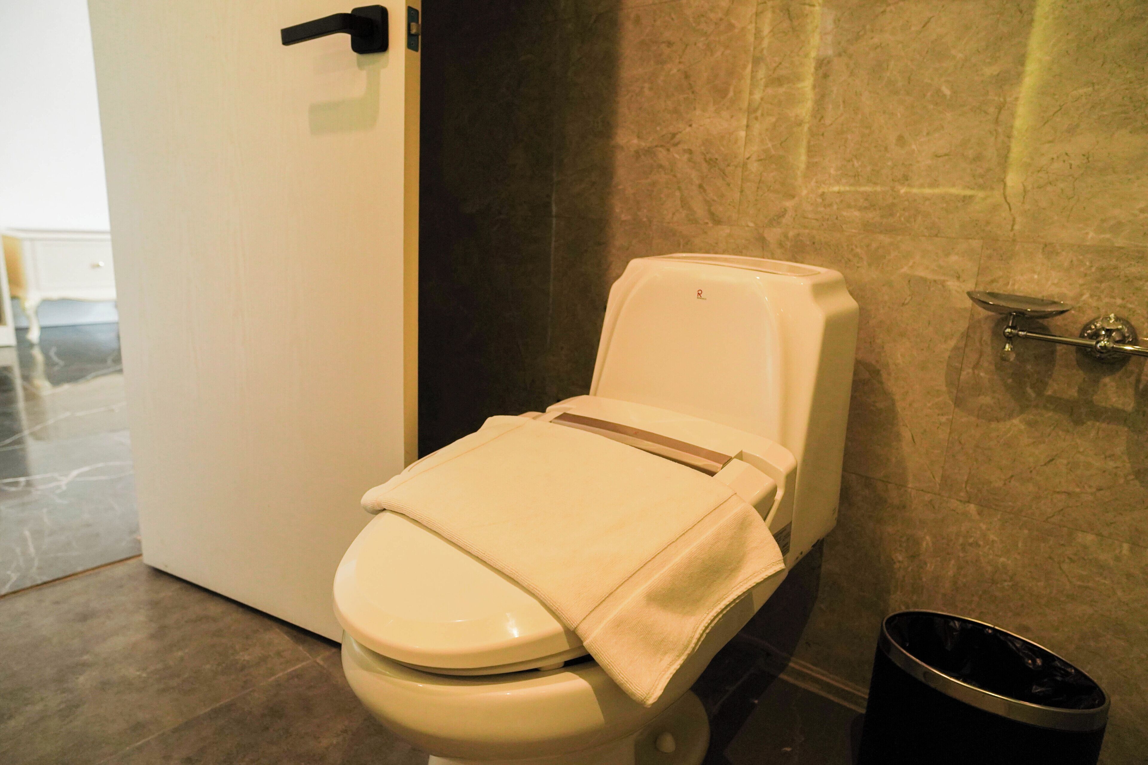 A toilet with a towel on the seat, a black trash bin, and a marble floor.