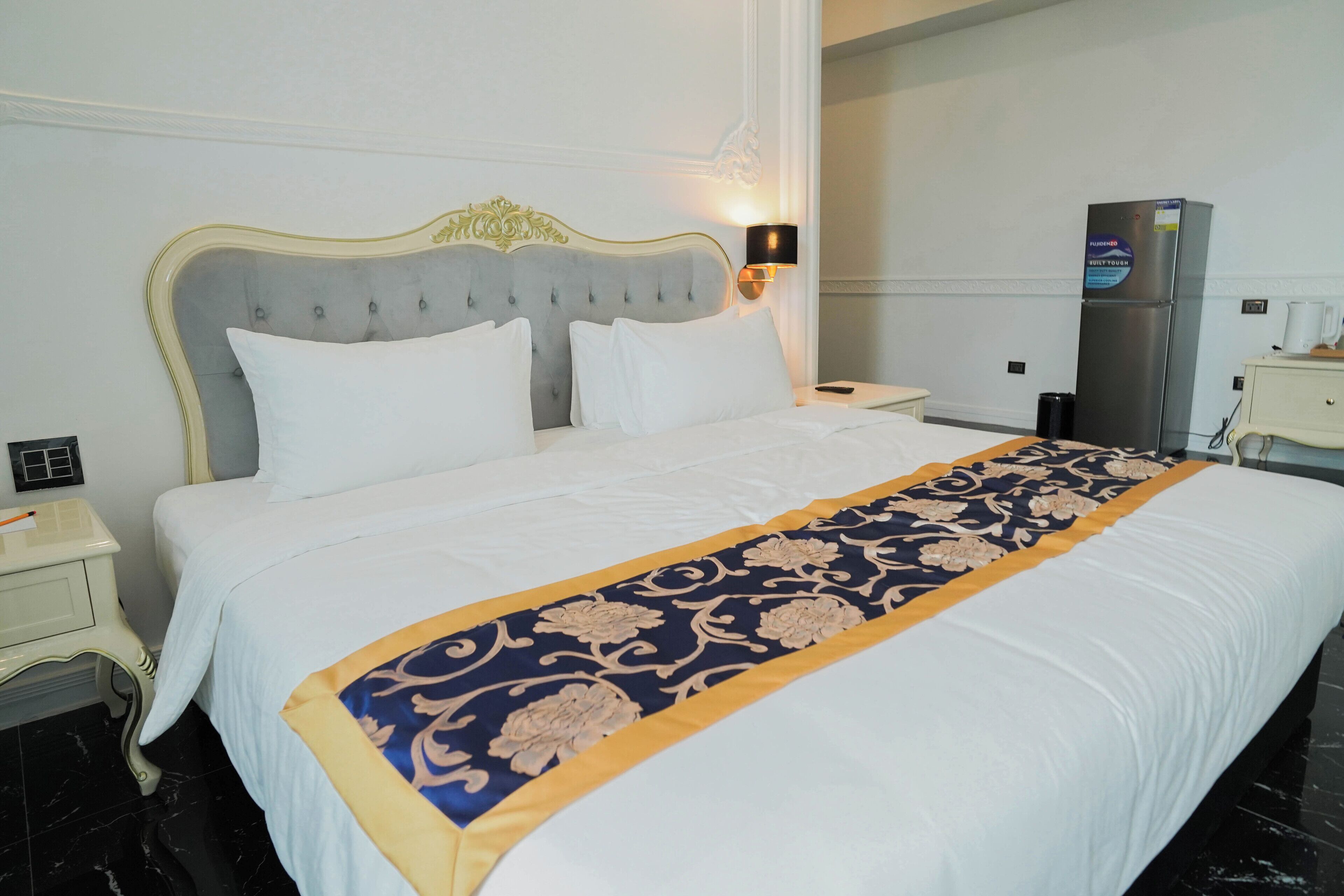 Habitación de hotel con una cama grande, una mesita de noche, una lámpara, un refrigerador y una pequeña mesa con una taza.