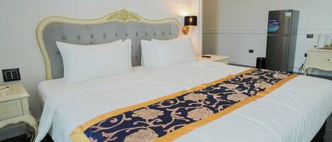 Habitación de hotel con una cama grande, una mesita de noche, una lámpara, un refrigerador y una pequeña mesa con una taza.