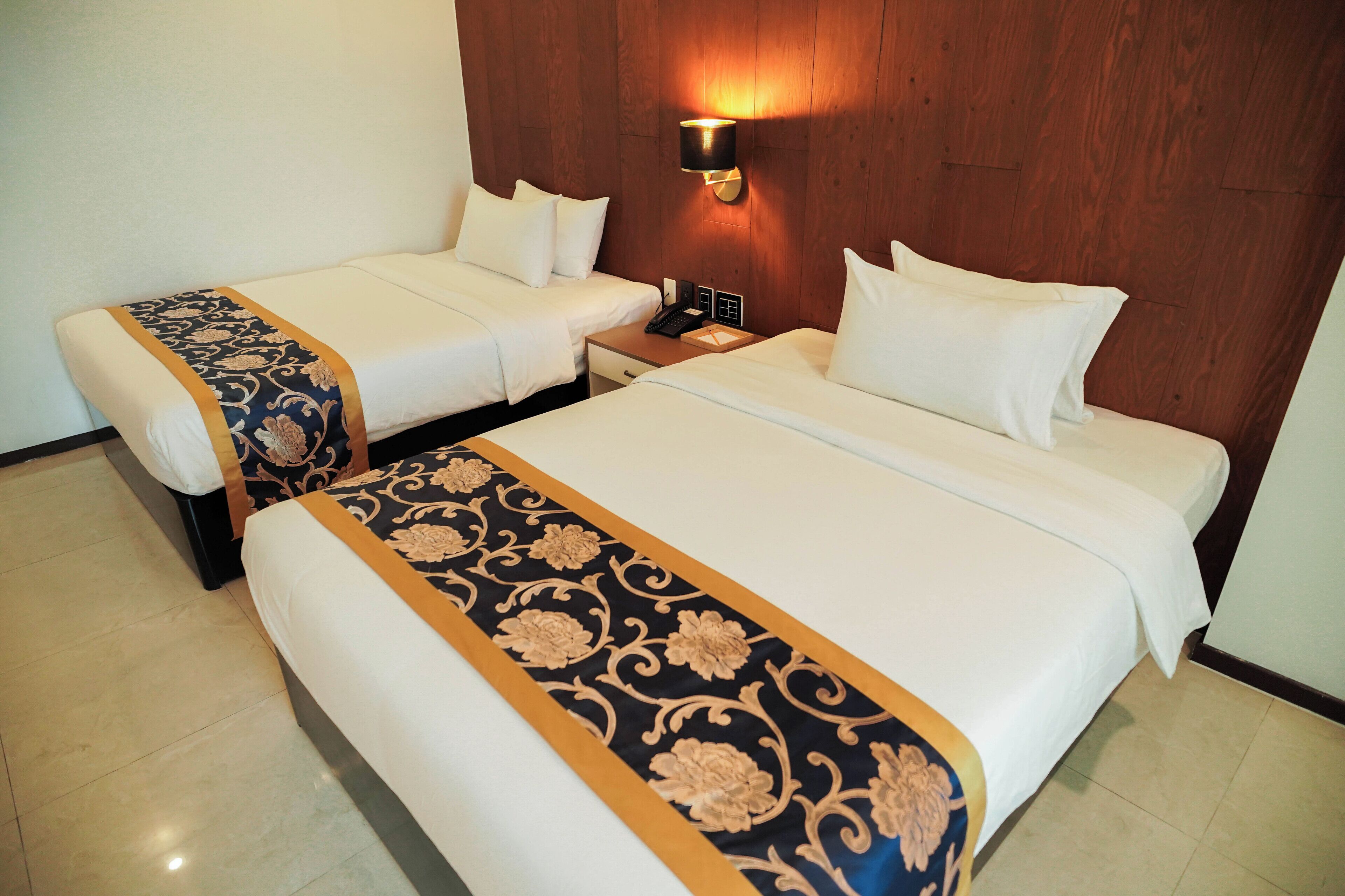 A hotel room with two beds, a nightstand, and a wall-mounted lamp.