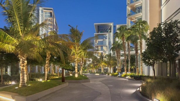 A modern hotel corridor with palm trees, a curved pathway, and contemporary buildings in the background.