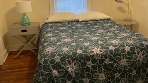 Un dormitorio con cama, mesita de noche, lámpara, ventana con persianas y dos cuadros colgados en la pared.