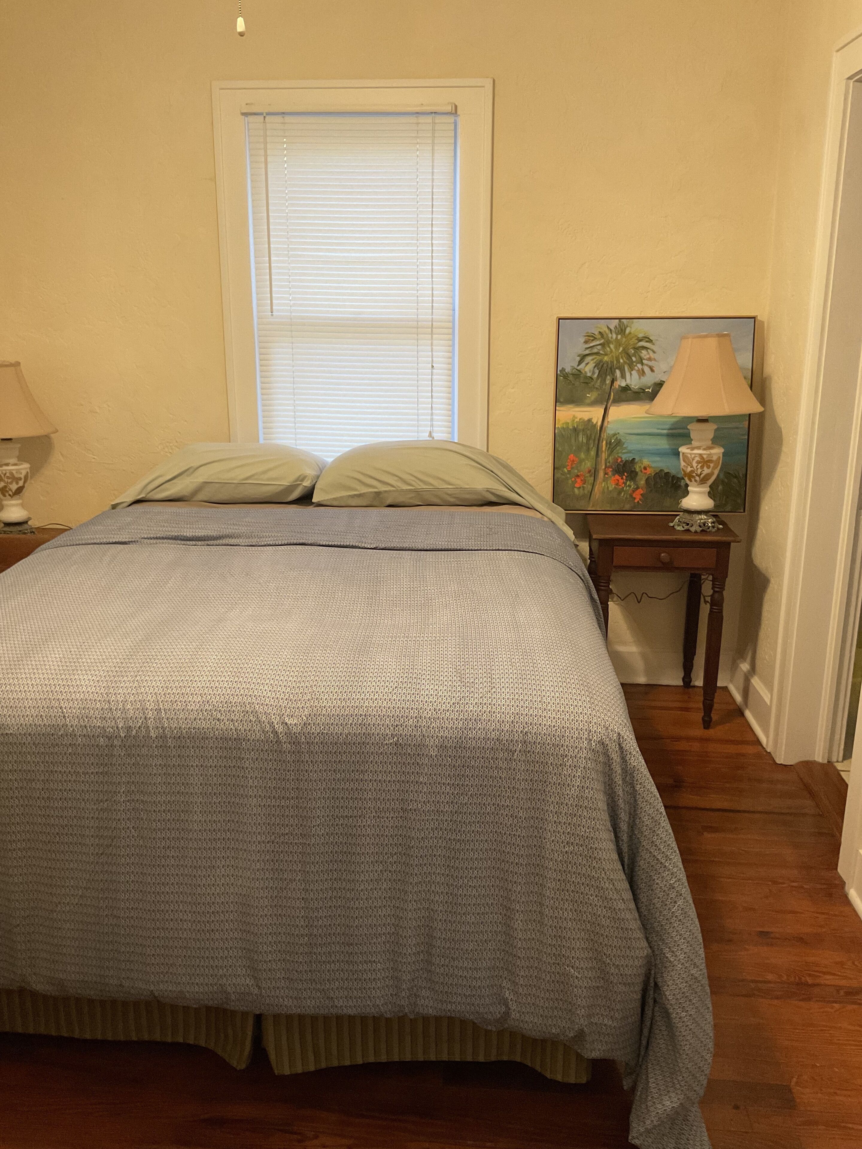 A bedroom with a bed, a nightstand with a lamp, a painting, and a window with blinds.