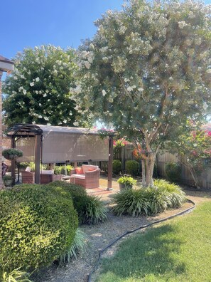 Un patio con zona de estar cubierta, rodeado de setos cuidados y diversas plantas.