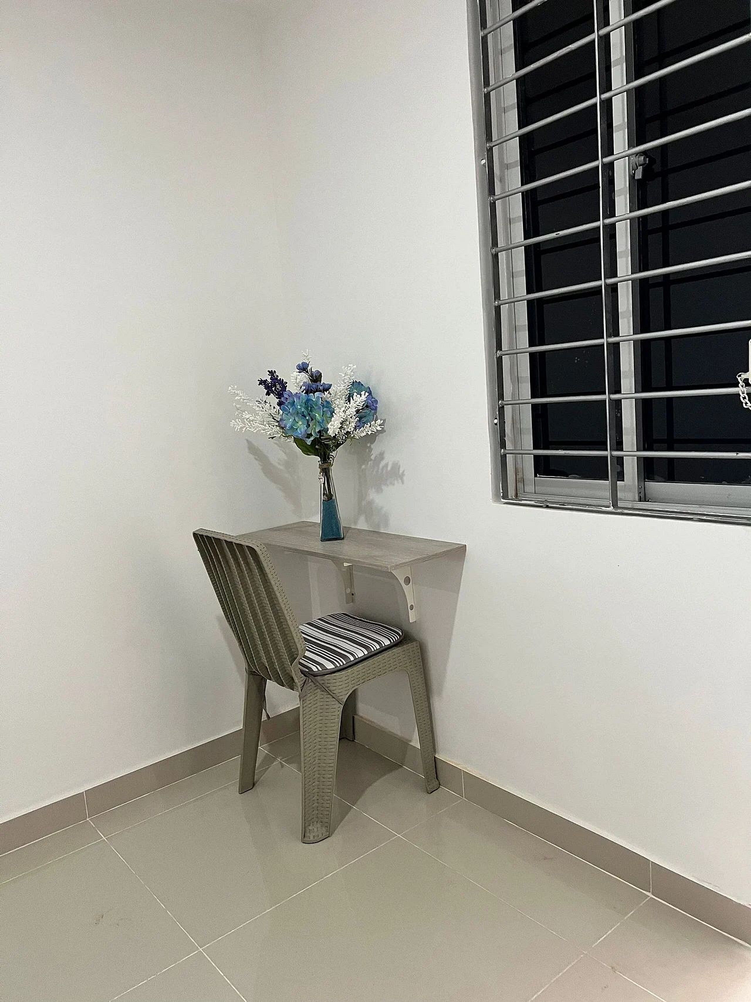A corner room with a chair, a small table, a vase with flowers, and a window with metal bars.