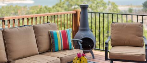 A deck with a sofa and armchair, a table with decorative items, and a view of a body of water and greenery.