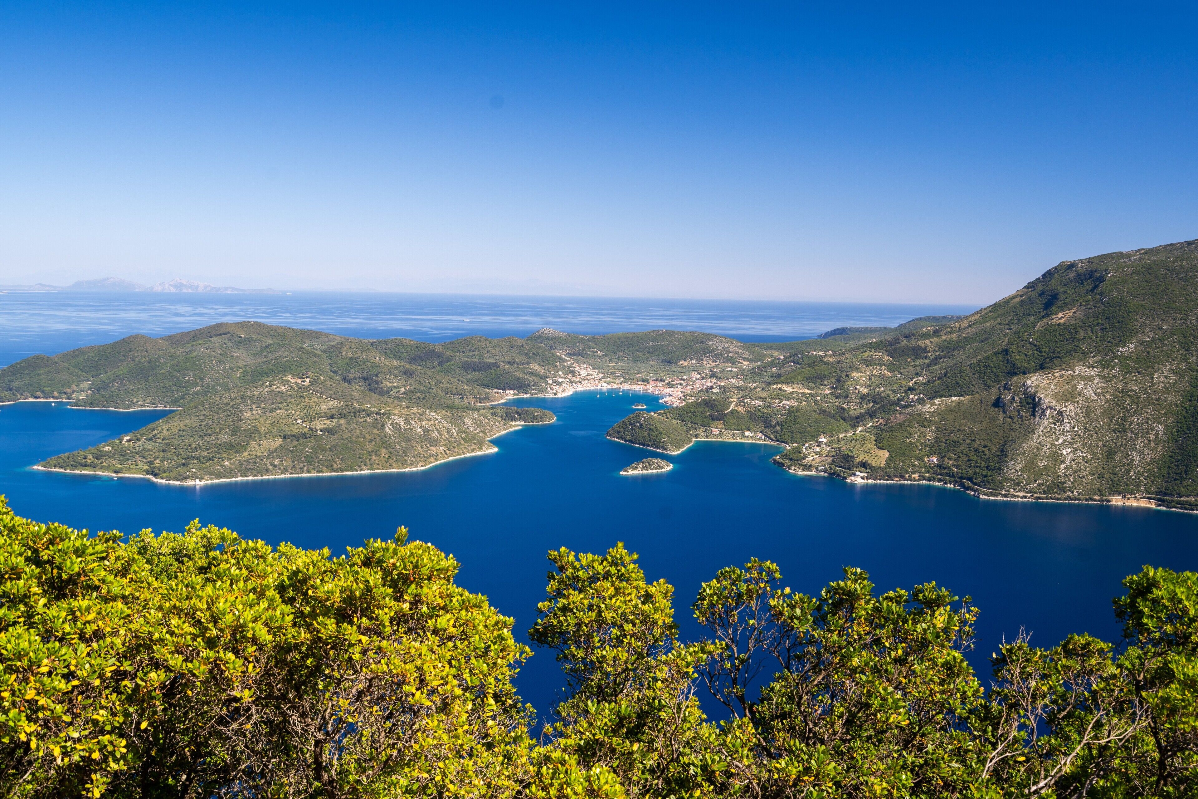 A coastal landscape with clear blue waters, green hills, and dense tree coverage.