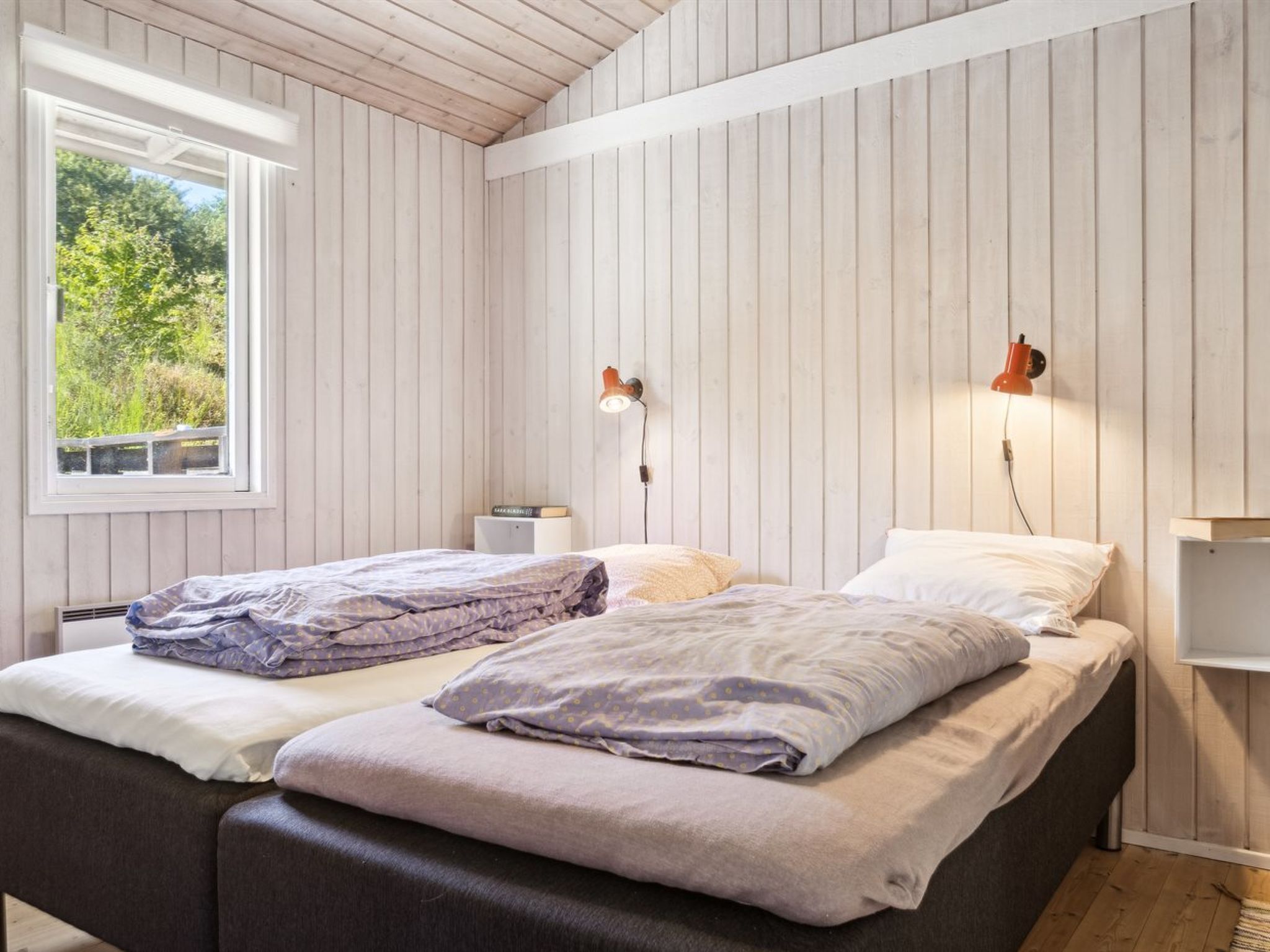 A bedroom with two beds, wooden panel walls, a window with a view of greenery, and two wall-mounted lamps.