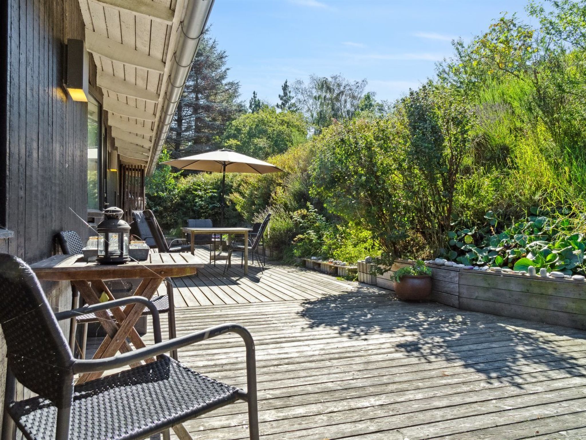 A wooden deck with outdoor furniture, including chairs and a table, surrounded by lush greenery and a stone wall.