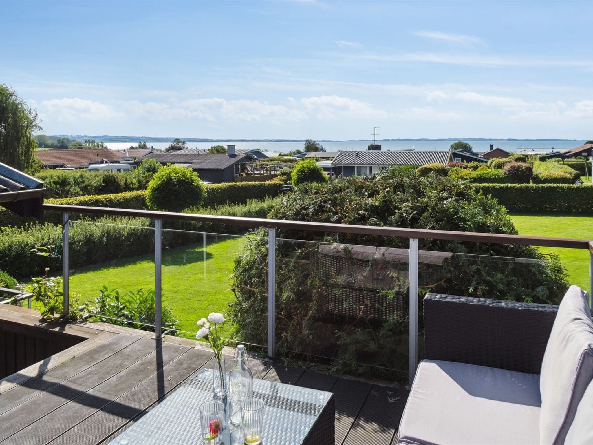 Une terrasse avec une table en verre, un canapé blanc et une vue sur un jardin aménagé et des bâtiments situés au loin.