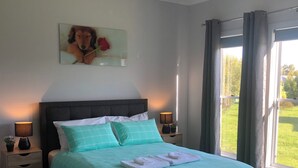 A neatly made bed with a teal bedspread and white pillows, two bedside lamps, a nightstand with a small plant, and a painting of a dog with a rose on the wall.