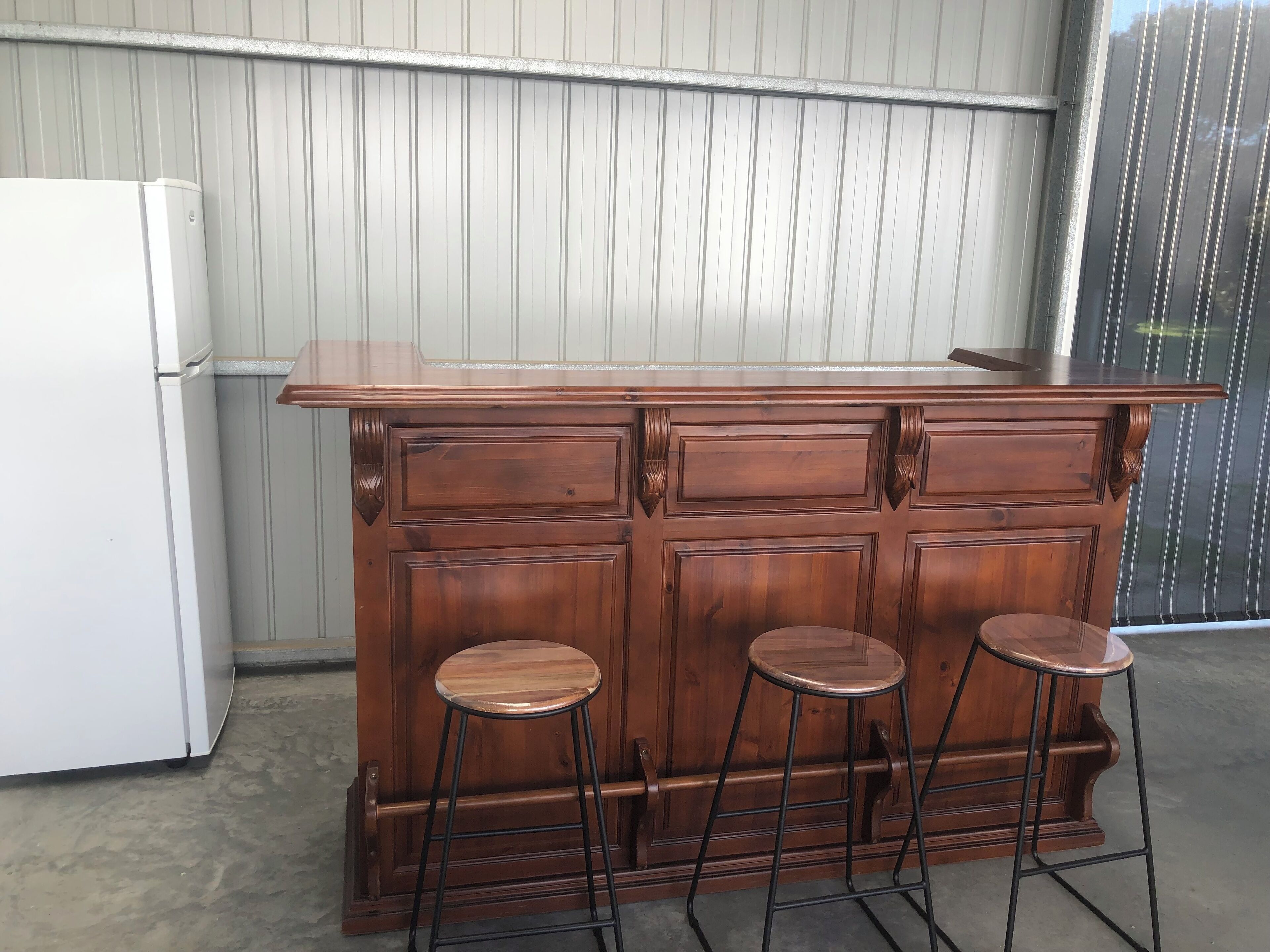 A wooden bar with three stools and a white refrigerator in a room with a corrugated metal wall.