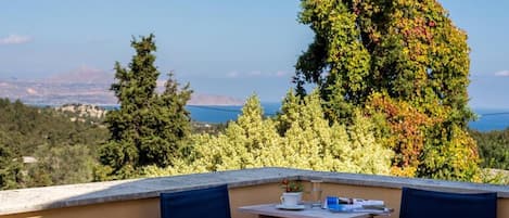 Terraço com duas cadeiras e uma mesa, com vista para uma paisagem deslumbrante com árvores e o mar.