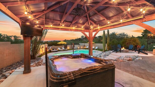 Un bain à remous sous une pergola en bois, illuminé de guirlandes lumineuses, entouré de chaises longues et d’une piscine.