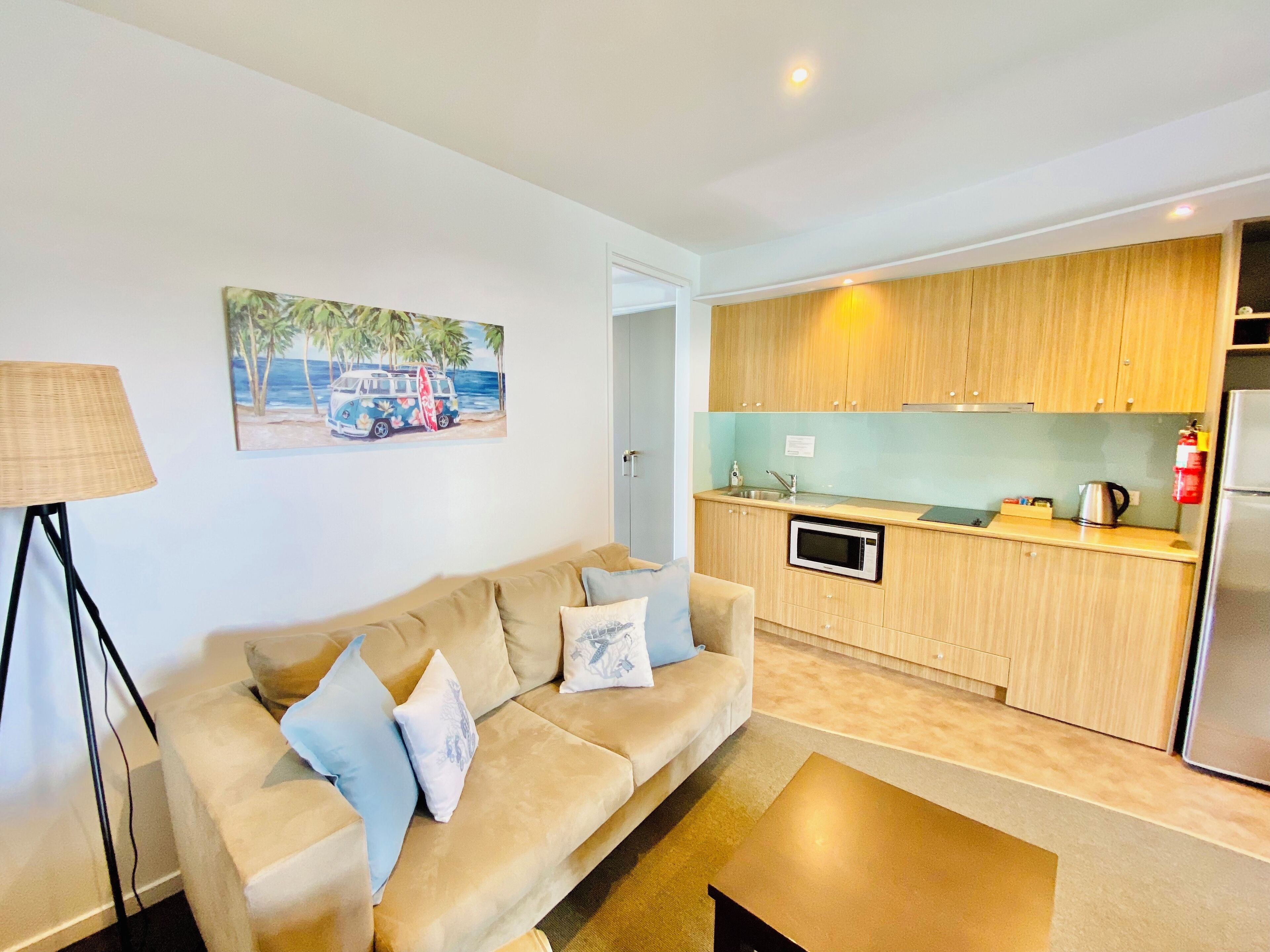 A modern living room with a beige sofa and a coffee table, a kitchen with wooden cabinets and a built-in microwave, and a painting of a beach scene on the wall.