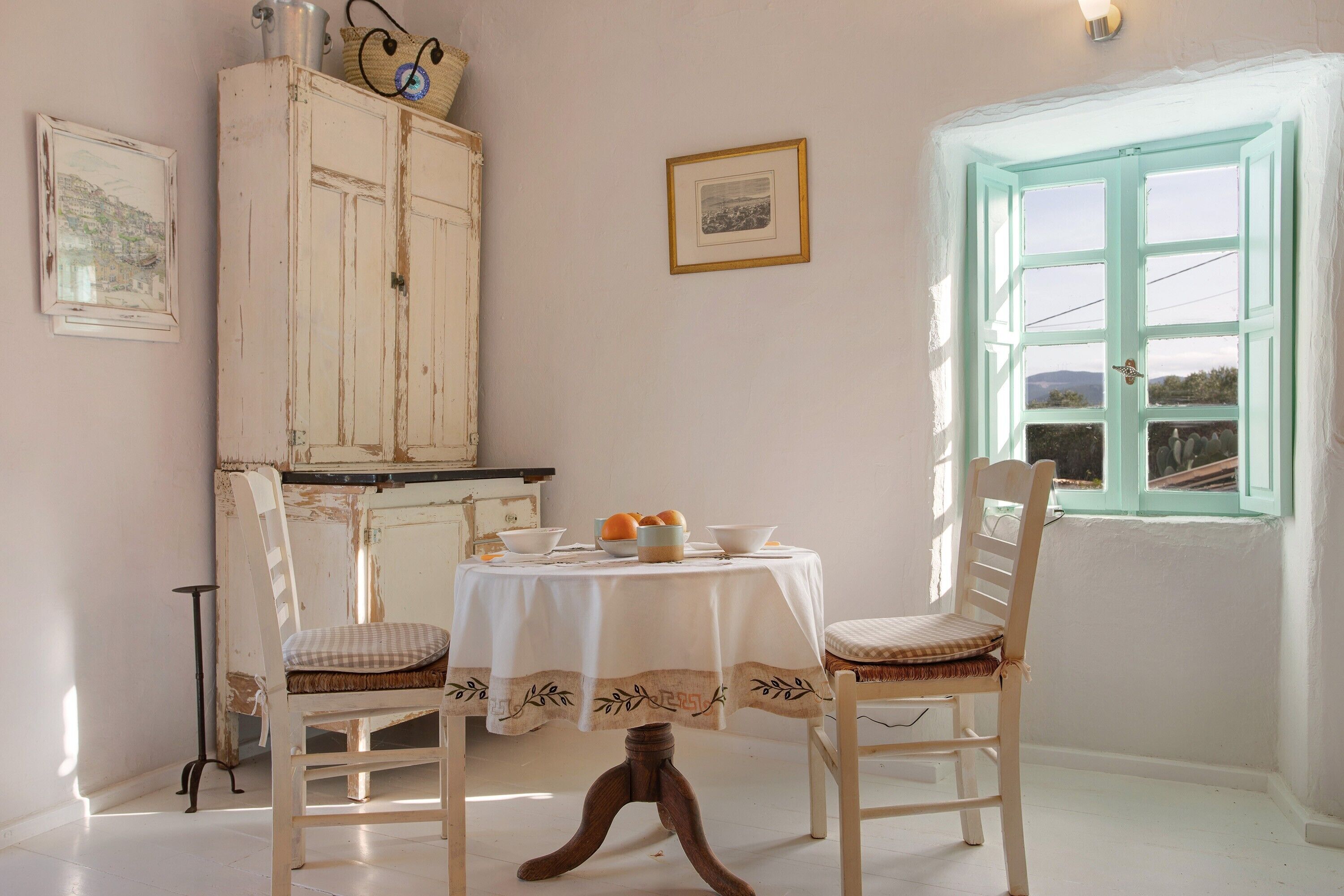 A room with a table set for two, a cabinet, a window with a view, and a framed picture on the wall.