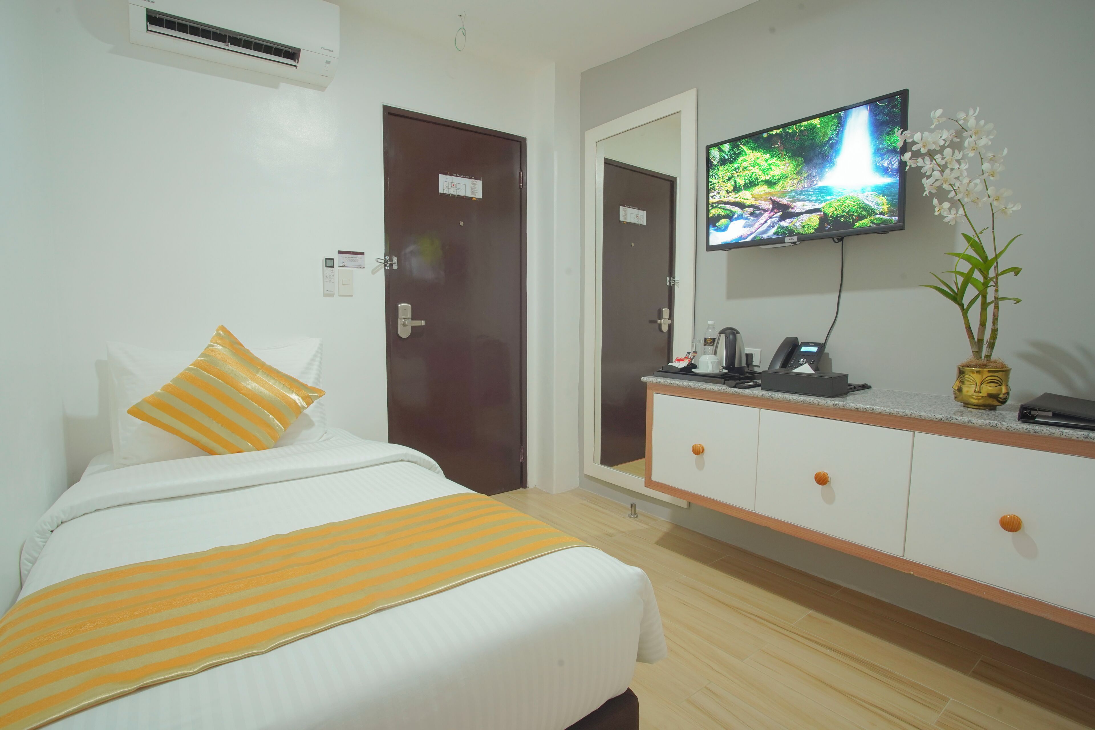 A hotel room with a bed, a television mounted on the wall, a mirror, and a small cabinet.