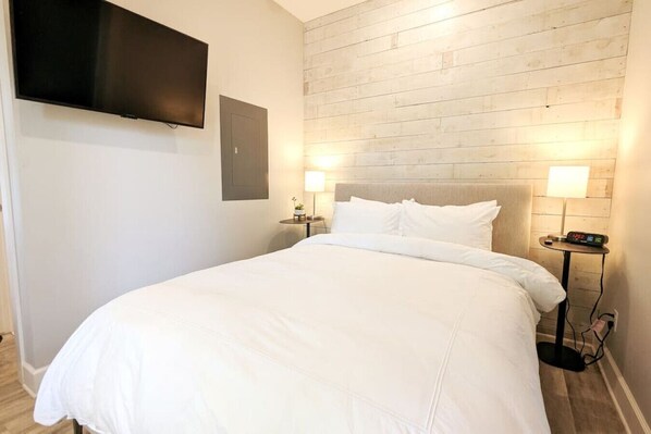 A hotel room with a large bed, two bedside tables with lamps, a flat-screen TV mounted on the wall, and a wooden headboard. - Downtown+HotTub+Wifi+FreeParking @151 Spring Ste B (Eureka Springs)